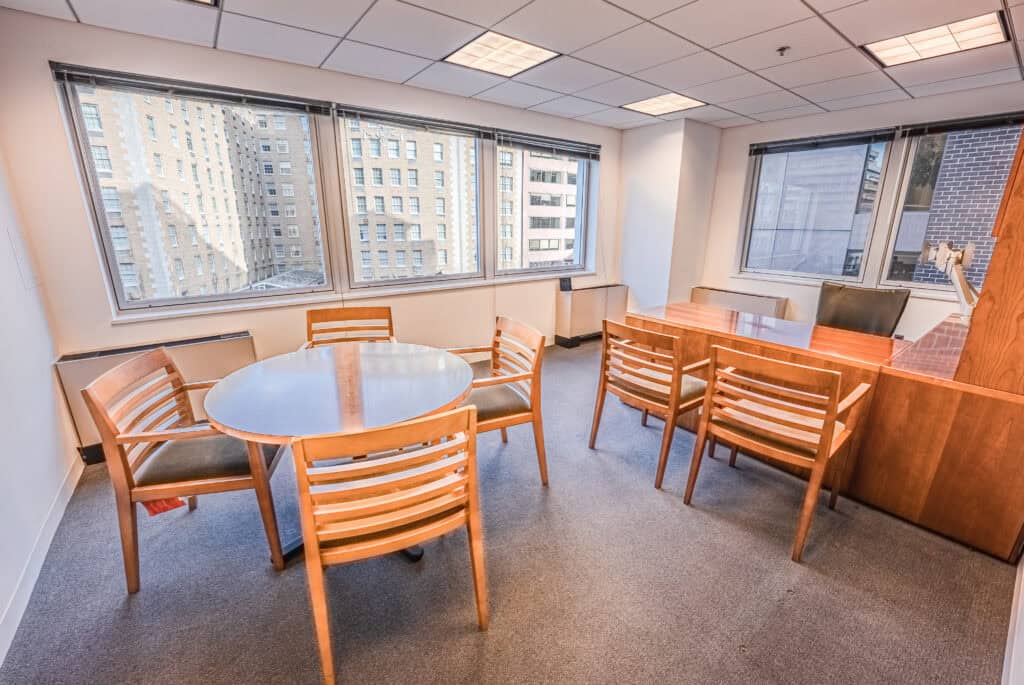 Bright private office with a round meeting table, wooden chairs, and a large executive desk in front of wide windows overlooking neighboring city buildings.