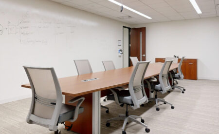 A brightly lit, modern conference room in Downers Grove IL with a long wooden table, gray mesh chairs, and a wall used as a large whiteboard covered in writing.