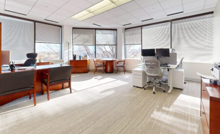 A large, bright corner office in Downers Grove IL with extensive windows covered by horizontal blinds, featuring a wooden executive desk and a separate modern white workstation with two monitors, plus a small circular meeting table.
