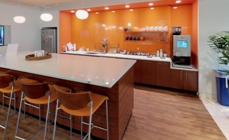 Modern office kitchen at Washington DC with a long white countertop island, orange bar stools, brown cabinets, a bright orange backsplash, and a coffee station.