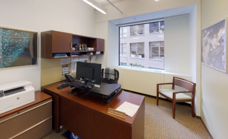 Private office in Washington DC with a dark wood desk and hutch, a printer on a matching cabinet, a monitor on a riser, and a wooden side chair near a window overlooking a brick building.