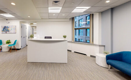 Office reception area in Boston MA with a white reception desk, a blue sofa, a tree painting, and two turquoise chairs, with a curved window looking out onto a city building.