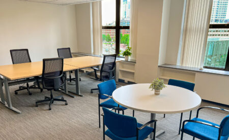 Small team office in Boston MA with a long light wood table and black mesh chairs, and a round white table with four blue upholstered chairs, next to large windows.